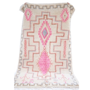 May include: A white Moroccan rug with a pink and gray diamond pattern. The rug has a fringe border.