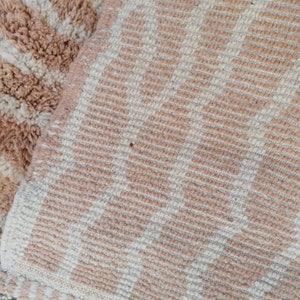 Puede incluir: Primer plano de una alfombra tejida a rayas beige y blancas con textura peluda. La alfombra tiene un sutil patr&oacute;n de l&iacute;neas diagonales.