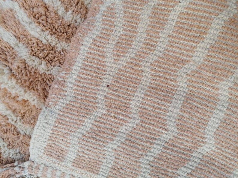 Puede incluir: Primer plano de una alfombra tejida a rayas beige y blancas con textura peluda. La alfombra tiene un sutil patr&oacute;n de l&iacute;neas diagonales.
