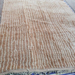 Puede incluir: Una alfombra tejida beige y blanca con un patr&oacute;n ondulado. La alfombra tiene un borde de flecos.