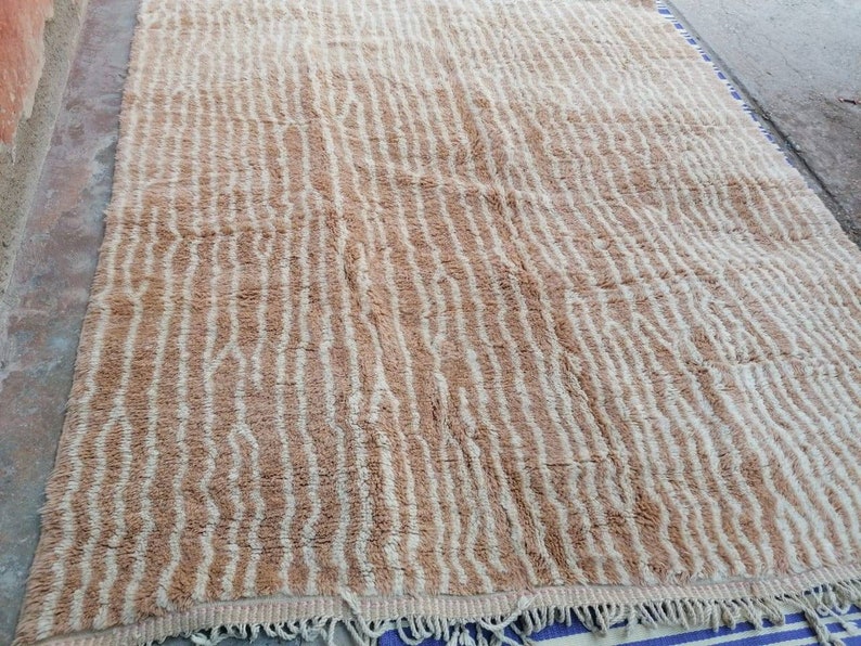 Puede incluir: Una alfombra tejida beige y blanca con un patr&oacute;n ondulado. La alfombra tiene un borde de flecos.