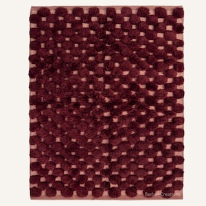 May include: A rectangular rug featuring a grid pattern of deep burgundy tufted pom-poms set against a light mauve base. The surface has a high-pile texture with repeating circular tufts. The text "Berber Creations" is visible in the bottom corner.