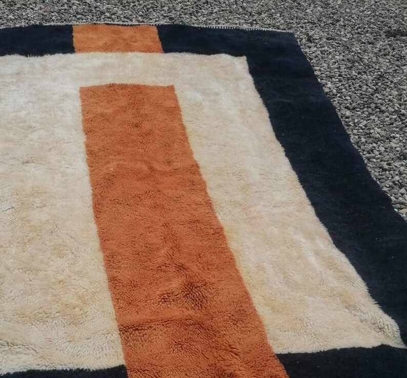 K&ouml;nnte beinhalten: Ein rechteckiger Teppich mit geometrischem Muster. Der Teppich hat einen schwarzen Rand, eine wei&szlig;e Mitte und einen braunen Streifen in der Mitte.