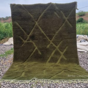 Pode incluir: Tapete verde-oliva com um padrão de losangos. O tapete tem uma superfície texturizada e uma borda com franjas. O tapete é exibido ao ar livre, com um fundo natural de grama e um céu nublado.