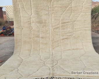 May include: A large, cream-colored rug with a geometric design. The rug has a thick, textured pile and a fringe along the bottom edge. The design features vertical and horizontal lines, creating a pattern. The rug is displayed outdoors.