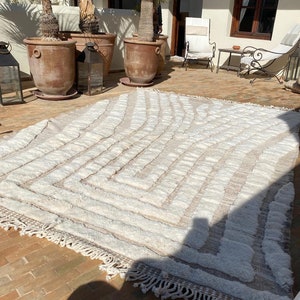 Puede incluir: Una alfombra tejida blanca y beige con un patrón geométrico y bordes con flecos. La alfombra está colocada en un patio de ladrillo.