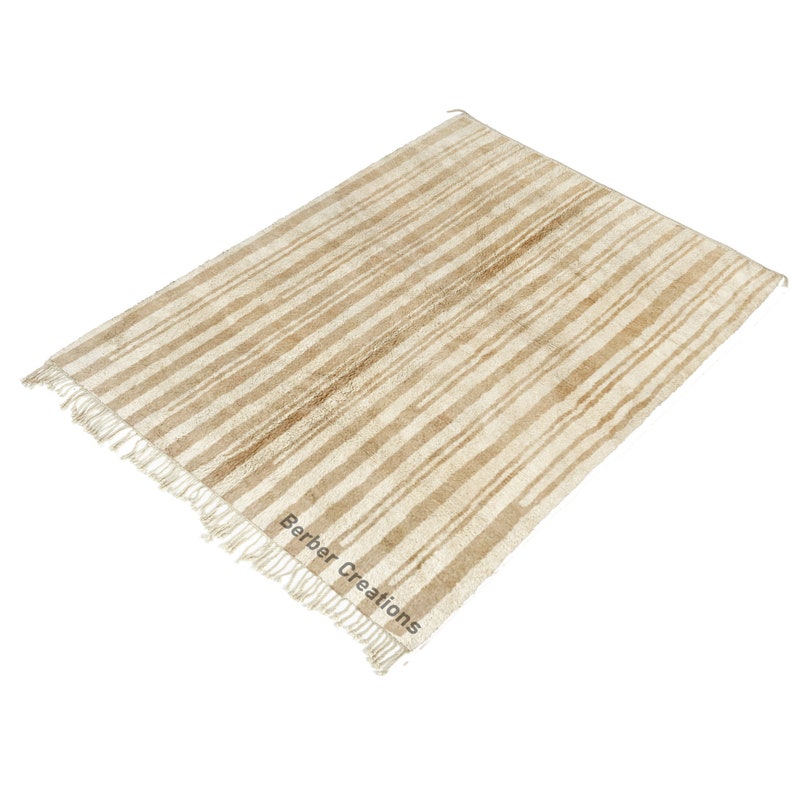 May include: A cream-colored rug with brown stripes and fringe. The rug has the text "Berber Creations" in the bottom right corner.