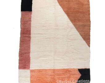 May include: A white, handwoven rug with geometric shapes in shades of pink, orange, and brown. The rug has a fringe border.