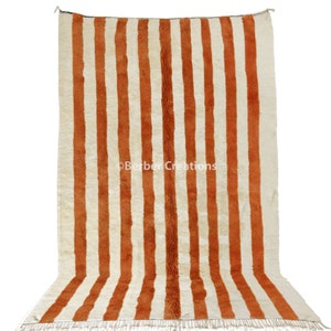 May include: A white and orange striped rug with a fringe border. The rug is made of wool and has a traditional Moroccan design.