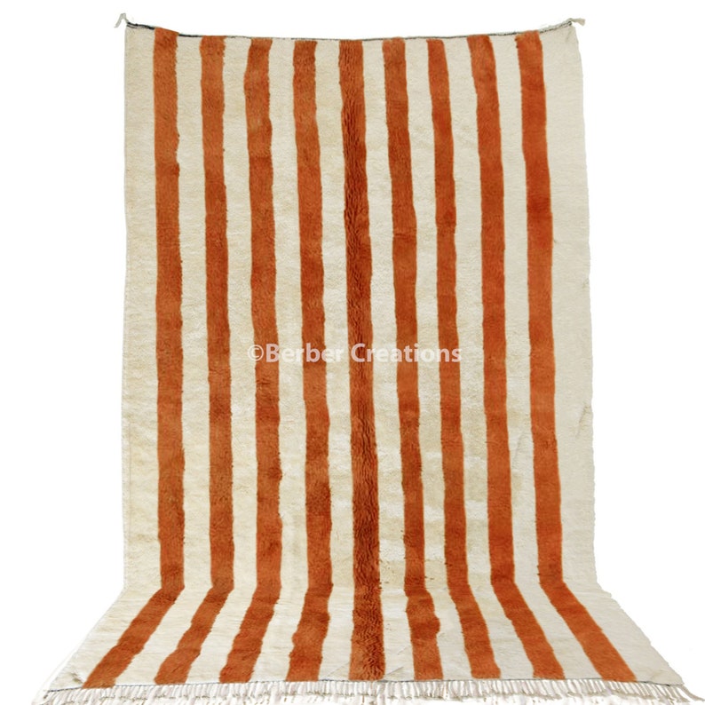May include: A white and orange striped rug with a fringe border. The rug is made of wool and has a traditional Moroccan design.