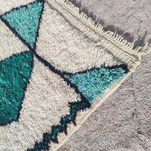 May include: Close-up of a white and green rug with a geometric pattern. The rug has a fringe border and is made of a soft, fluffy material.