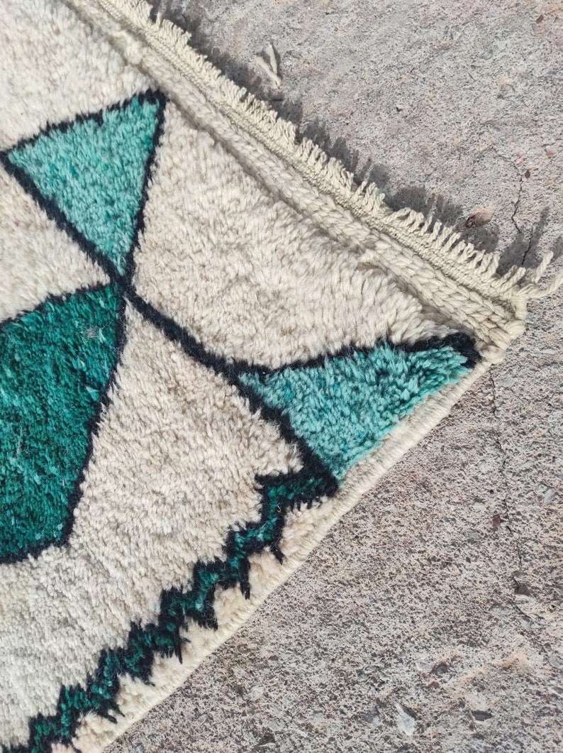 May include: Close-up of a white and green rug with a geometric pattern. The rug has a fringe border and is made of a soft, fluffy material.