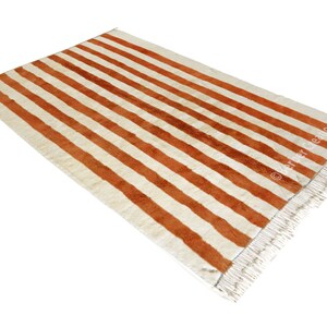 May include: A white and orange striped rug with fringe on the edges. The rug has a woven texture.
