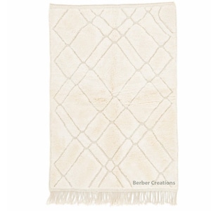 May include: A cream-colored rug with a diamond pattern woven into the fabric. The rug has fringe along the bottom edge and the text "Berber Creations" is printed in the bottom right corner.