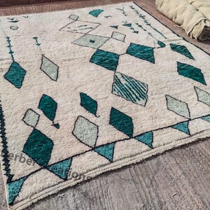 May include: A white rug with a geometric pattern of teal and green diamonds. The rug has a fringe border.