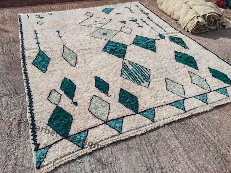 May include: A white rug with a geometric pattern of teal and green diamonds. The rug has a fringe border.