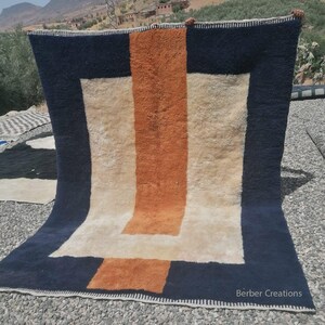 K&ouml;nnte beinhalten: Ein handgewebter Teppich mit einem geometrischen Muster in Wei&szlig;-, Orange- und Marineblau-T&ouml;nen. Der Teppich hat einen Fransenrand und der Text "Berber Creations" ist in der rechten unteren Ecke sichtbar.