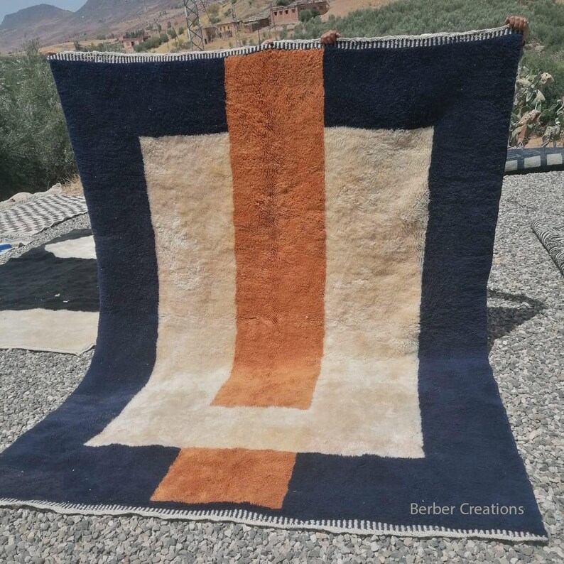 K&ouml;nnte beinhalten: Ein handgewebter Teppich mit einem geometrischen Muster in Wei&szlig;-, Orange- und Marineblau-T&ouml;nen. Der Teppich hat einen Fransenrand und der Text "Berber Creations" ist in der rechten unteren Ecke sichtbar.
