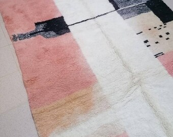 May include: A hand-woven rug with a geometric pattern in shades of pink, white, black, and gray. The rug has a fringe border.