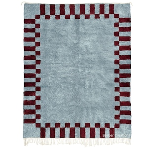 May include: A rectangular rug with a light blue center and a dark red and light red checkered border. The rug has a shaggy texture and white fringe along the bottom edge. The words "Berber Creations" are printed in the bottom right corner.
