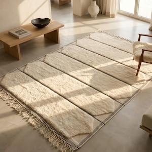 May include: A rectangular off-white wool rug featuring a geometric pattern of elongated hexagonal shapes outlined in dark brown thread. The rug has a thick, plush texture and is finished with long, knotted fringe tassels along the short edges.