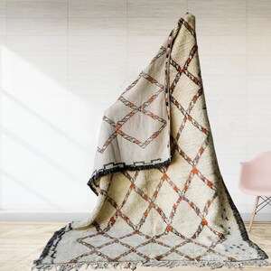 May include: A white and orange Moroccan rug with a diamond pattern. The rug is folded and draped over a chair in a room with white walls and a wooden floor.