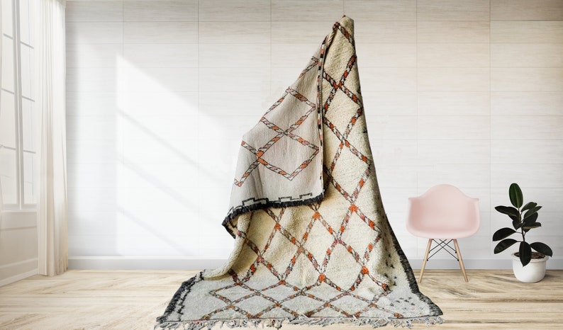 May include: A white and orange Moroccan rug with a diamond pattern. The rug is folded and draped over a chair in a room with white walls and a wooden floor.