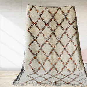 May include: A white and orange Moroccan rug with a diamond pattern. The rug has a fringe border and is laid out on a wooden floor.