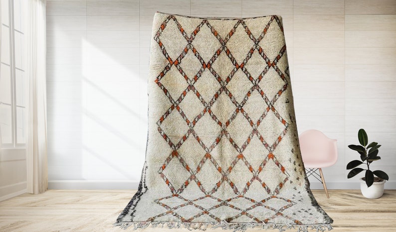 May include: A white and orange Moroccan rug with a diamond pattern. The rug has a fringe border and is laid out on a wooden floor.
