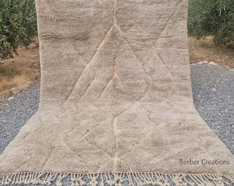 Puede incluir: Una alfombra marroquí grande, de color blanco roto, tejida a mano con un patrón geométrico. La alfombra tiene un borde de flecos y está hecha de lana. La alfombra se muestra sobre una superficie de grava.