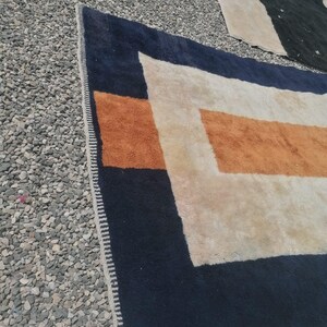 K&ouml;nnte beinhalten: Ein wei&szlig;er Teppich mit einem geometrischen Muster aus orangefarbenen und dunkelblauen Streifen. Der Teppich hat einen Fransenrand.