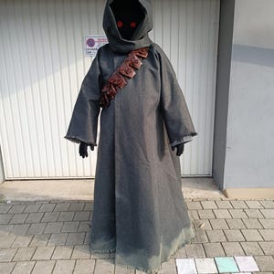 以下が含まれることがあります： 裾がほつれた長袖のダークグレーのフード付きローブ。胸元には複数の小さなポーチが付いた茶色のレザー製バンダリアが斜めに掛けられている。深く暗いフードの奥には、2つの光る赤い円形の目が見える。