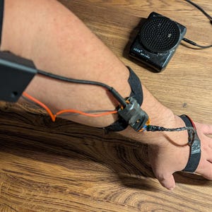 May include: A black electronic device with a rectangular battery pack strapped to the upper arm, connected by wires to a sensor module on the forearm and a wrist strap, with a separate black circular speaker unit positioned nearby.