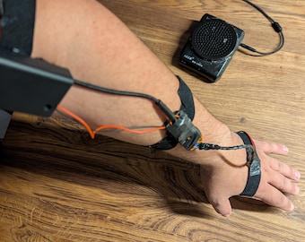 May include: A black electronic device with a rectangular battery pack strapped to the upper arm, connected by wires to a sensor module on the forearm and a wrist strap, with a separate black circular speaker unit positioned nearby.
