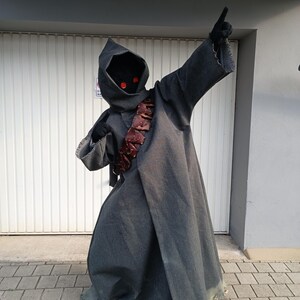May include: A full-length dark gray hooded robe costume featuring frayed edges at the sleeves and hem. The outfit includes a dark face covering with two glowing red circular eyes and a brown leather-like bandolier worn diagonally across the chest.