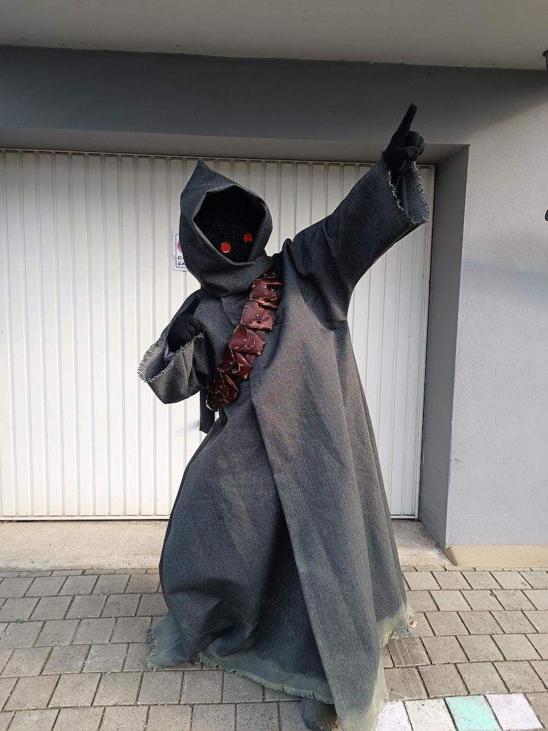 May include: A full-length dark gray hooded robe costume featuring frayed edges at the sleeves and hem. The outfit includes a dark face covering with two glowing red circular eyes and a brown leather-like bandolier worn diagonally across the chest.
