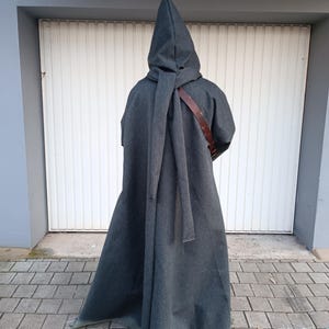 May include: A floor-length charcoal gray hooded cloak featuring a pointed hood and a long fabric tie draped down the back. A brown leather strap is visible across the shoulder area, worn over the garment, which extends to the ground.