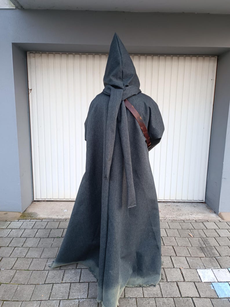 May include: A floor-length charcoal gray hooded cloak featuring a pointed hood and a long fabric tie draped down the back. A brown leather strap is visible across the shoulder area, worn over the garment, which extends to the ground.