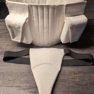 May include: A white fabric corset with two pockets on each side and a matching white fabric crotch strap with black elastic straps.