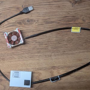 Könnte beinhalten: Ein kleiner brauner und silberner Computer-Kühlerlüfter, der mit einem schwarzen USB-Kabel und einem rechteckigen weißen Steuermodul verbunden ist. Das Kabel verfügt über ein gelbes Warnetikett und einen kleinen schwarzen Anschluss.