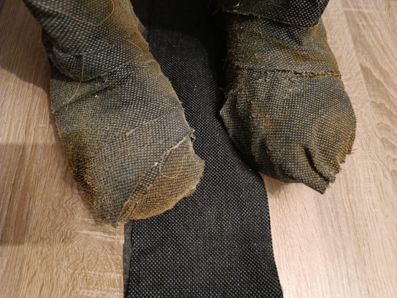 May include: A pair of dark, textured, fabric-covered boots with a worn, brownish-grey appearance and visible frayed edges. The boots feature a dense, woven material construction with a distinct grid-like pattern, showing signs of heavy use and wear.