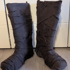 Puede incluir: Un par de botas de tela marrón con un aspecto desgastado. Las botas están hechas de una tela rugosa y texturizada y tienen una punta puntiaguda.