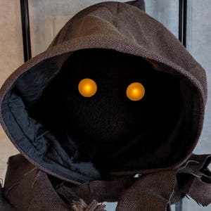 Pode incluir: Uma figura encapuzada marrom com dois olhos amarelos brilhantes. A figura é feita de tecido e tem uma aparência desfiada.