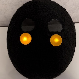 May include: A black plush toy with two yellow light-up eyes. The toy is shaped like a sphere.