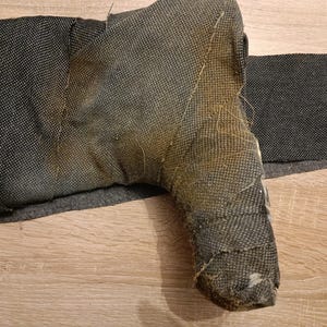 May include: A dark gray, textured, boot-shaped object wrapped in a woven fabric material. The surface shows a distinct transition from a dark charcoal color to a faded, brownish-yellow hue across the heel and ankle area, with visible frayed edges.
