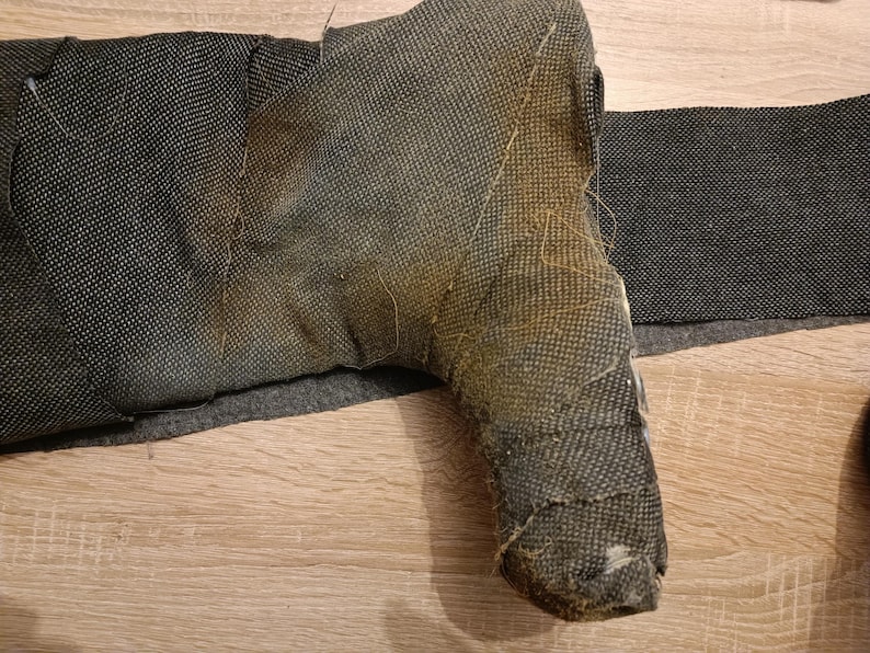 May include: A dark gray, textured, boot-shaped object wrapped in a woven fabric material. The surface shows a distinct transition from a dark charcoal color to a faded, brownish-yellow hue across the heel and ankle area, with visible frayed edges.