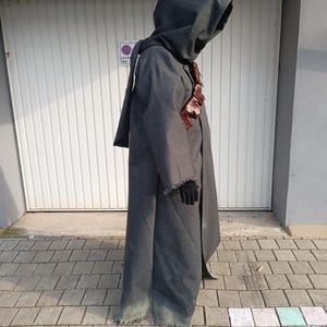 May include: A full-length, dark gray hooded cloak made of a textured fabric with a frayed hemline. The garment features a brown leather harness worn across the chest and is paired with black gloves. The hood is deep and covers the head area.