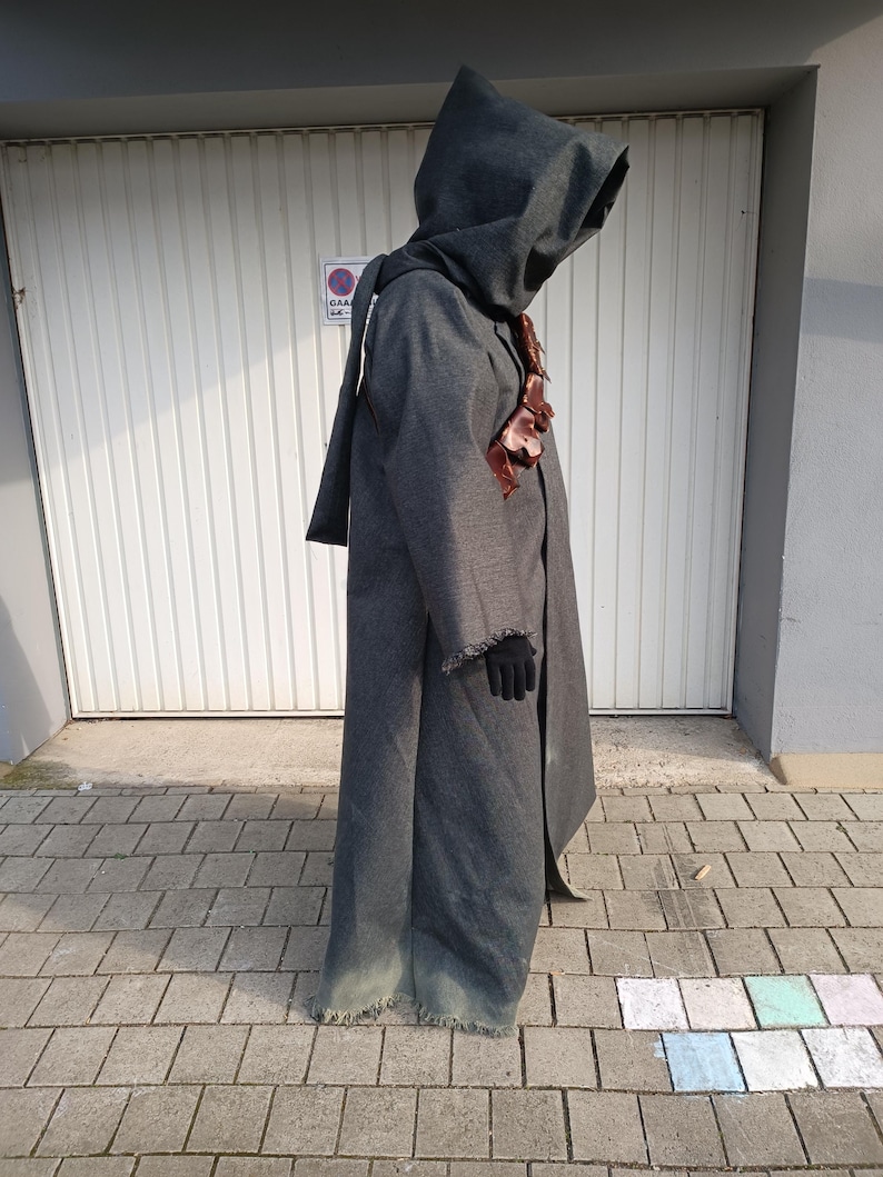 May include: A full-length, dark gray hooded cloak made of a textured fabric with a frayed hemline. The garment features a brown leather harness worn across the chest and is paired with black gloves. The hood is deep and covers the head area.