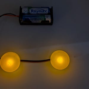 May include: A basic electrical circuit featuring two illuminated yellow lights linked to a battery holder containing two Raymax batteries. The wires are black and red, set against a grey backdrop.