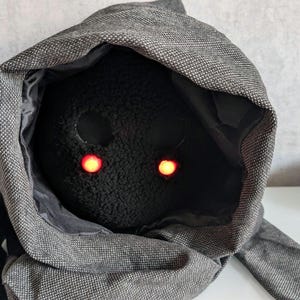 Könnte beinhalten: Ein Plüschtier mit einem schwarzen Kapuzenstoff, der seinen Kopf bedeckt. Das Spielzeug hat zwei leuchtende rote Augen.